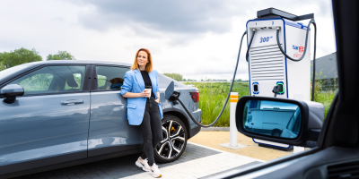 Vrouw leunt tegen haar auto tijdens het laden met een beker in haar hand.